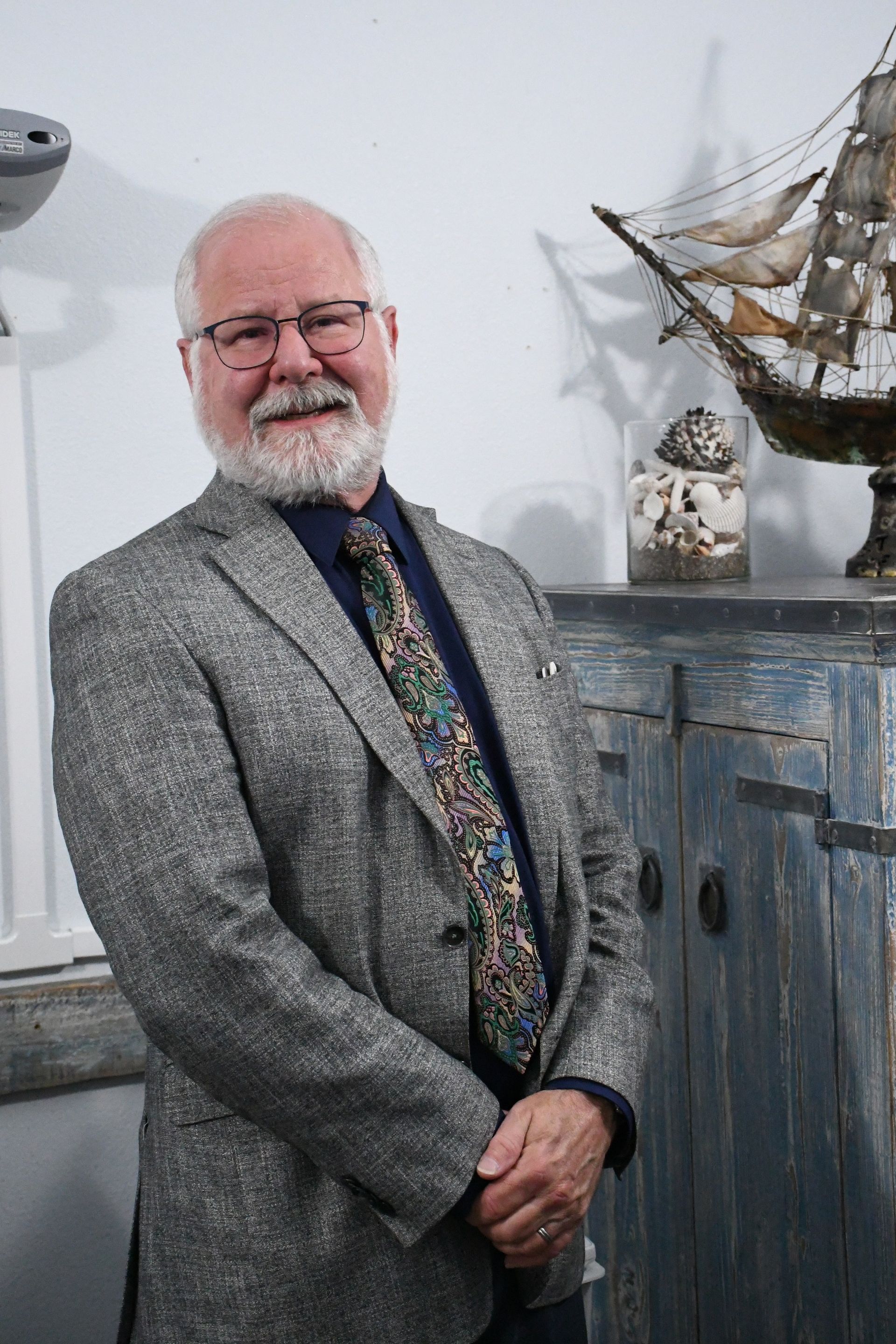 Dr. John Prewett, optometrist, standing with his hands clasped beside a blue cabinet and a model ship